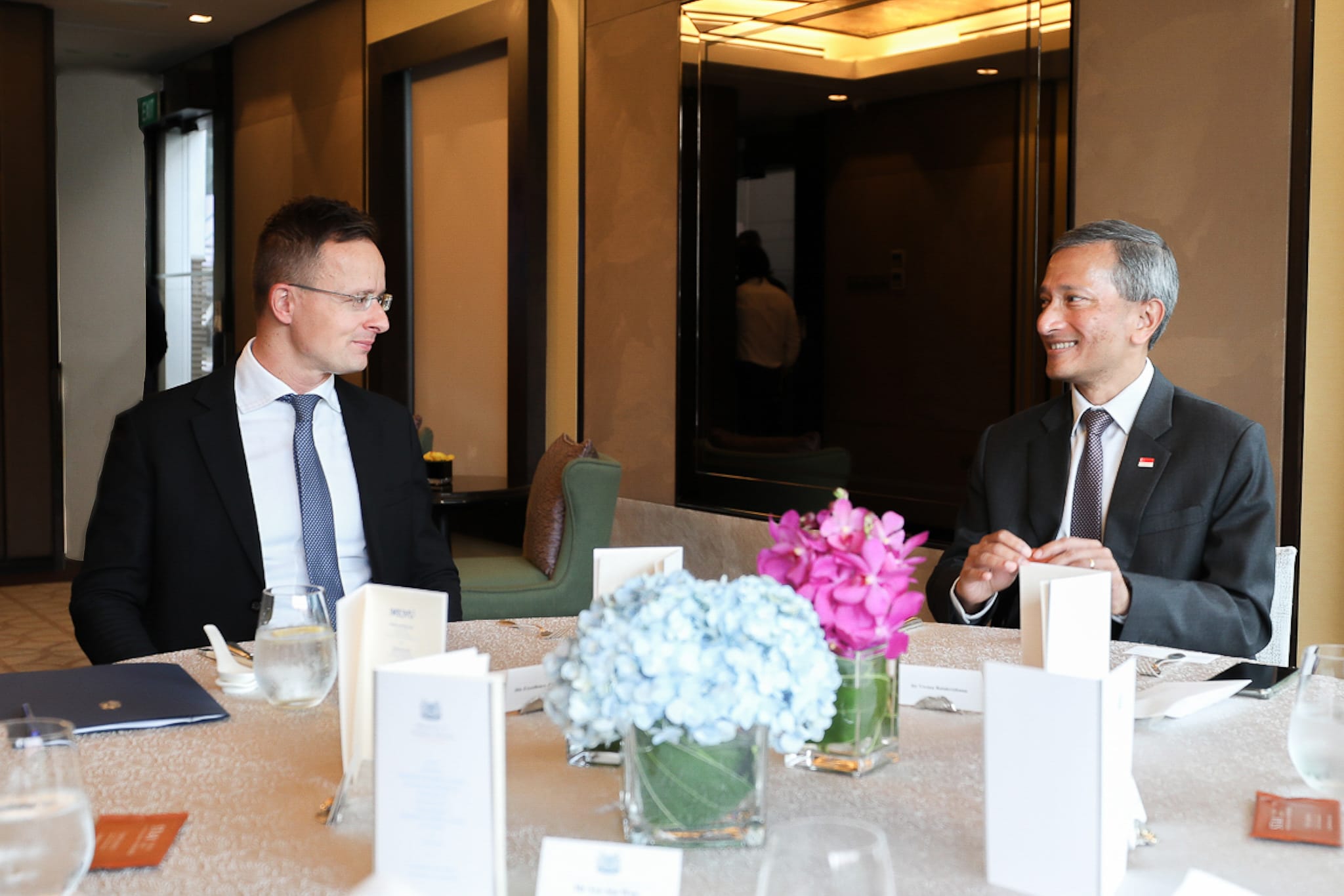 Two men in suits sit at a table with flowers, water glasses, and printed materials.